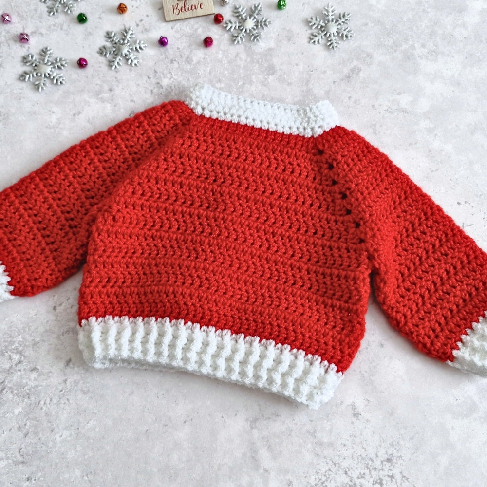 A red and white crocheted baby sweater with long sleeves, laid flat on a light gray textured surface with holiday decorations like silver snowflakes and colorful beads.