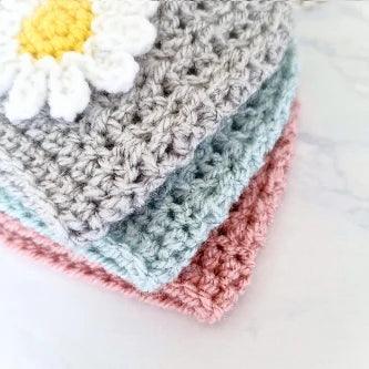 Close-up of textured crochet fabric squares in pink, blue, and gray with a white and yellow daisy applique