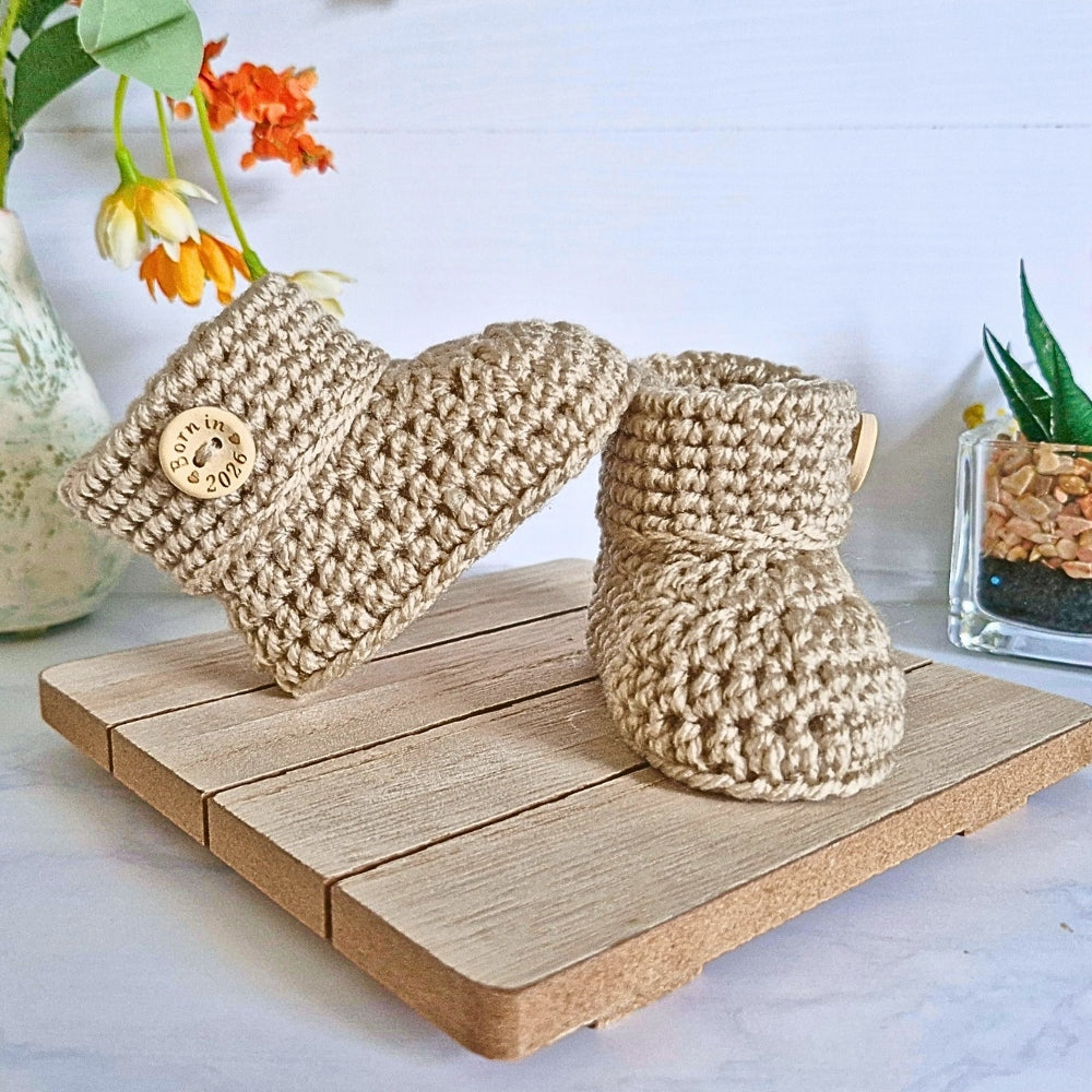 Pair of beige knitted baby boots on a wooden surface with plants in the background