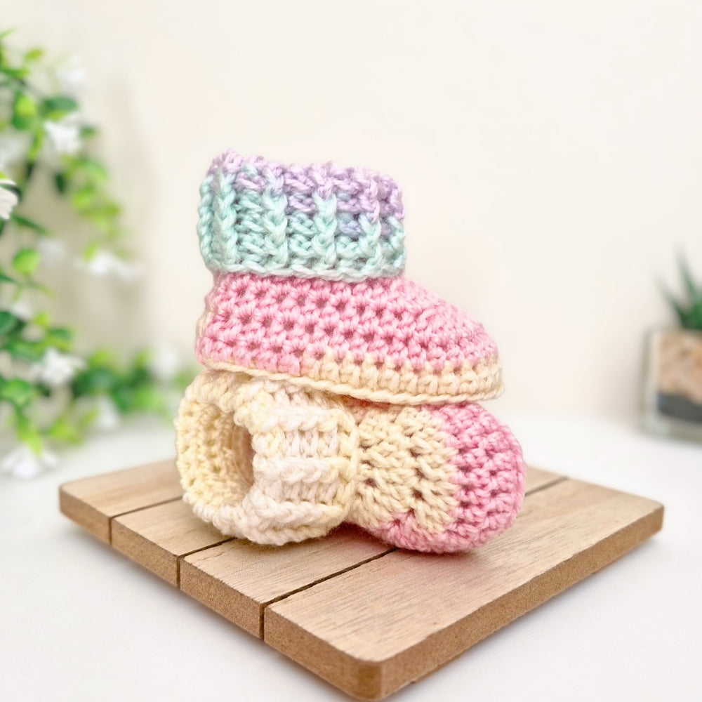 A pair of pastel crocheted booties stacked on a small wooden block, with a soft-focus indoor background and greenery.