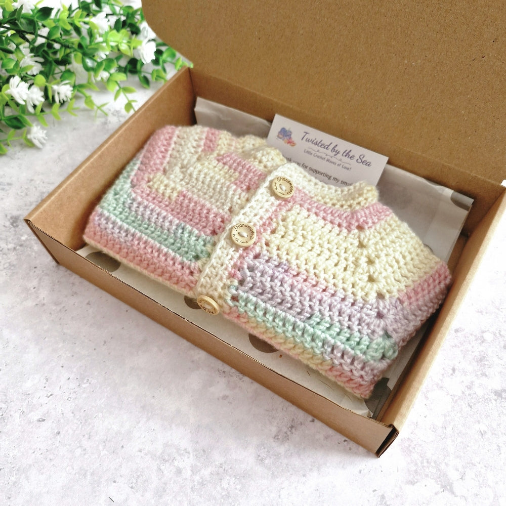 A pastel striped crocheted cardigan or sweater displayed inside an open brown cardboard box on a light surface, with a small card reading 'Twisted by the Sea' visible inside the box.