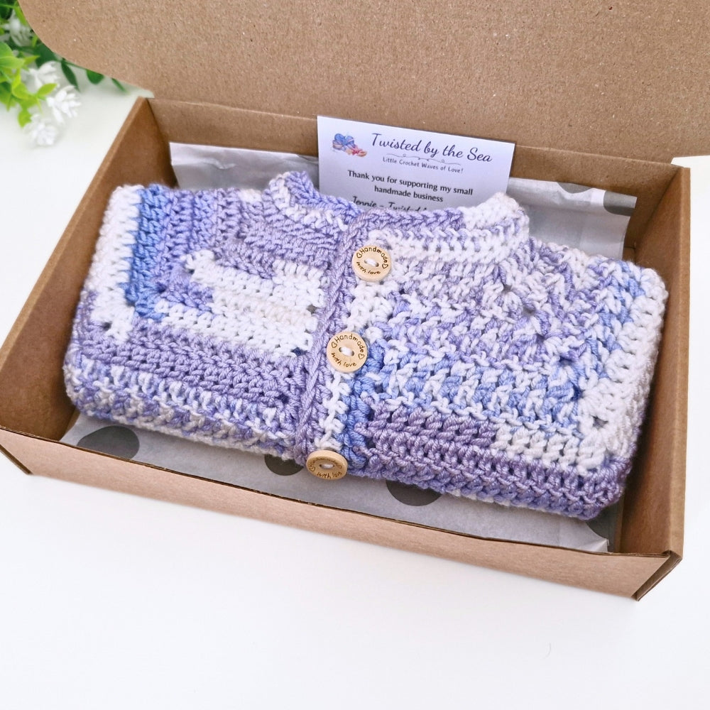 A handmade crocheted baby cardigan in lavender, light blue, and white, presented inside an open brown cardboard gift box with a branded card insert.