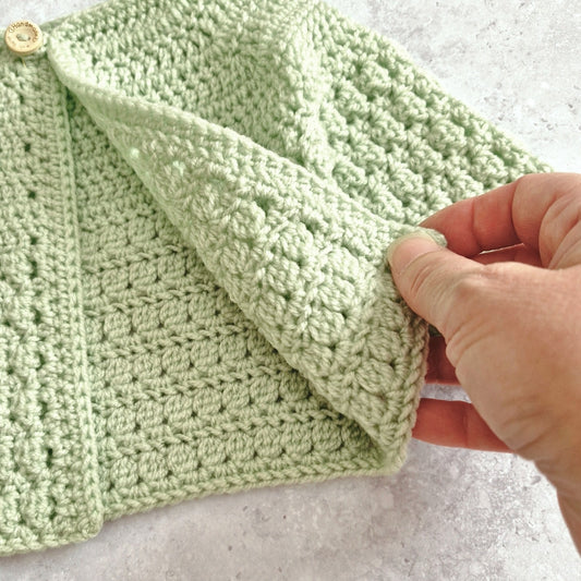 A close-up of a light sage green crocheted textile, likely a baby blanket or scarf, being held by a hand to show the texture. The item features a textured bobble stitch pattern, a ribbed border, and a small wooden button on the left, set against a light gray stone background.
