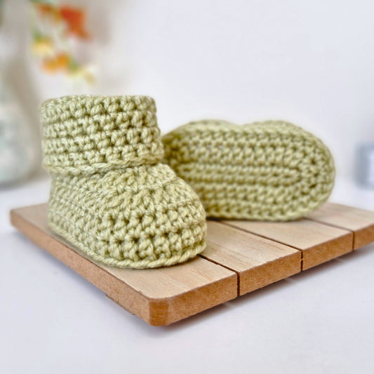 A Pair of newborn mushy peas green crochet baby booties on a wooden surface. Made with acrylic yarn