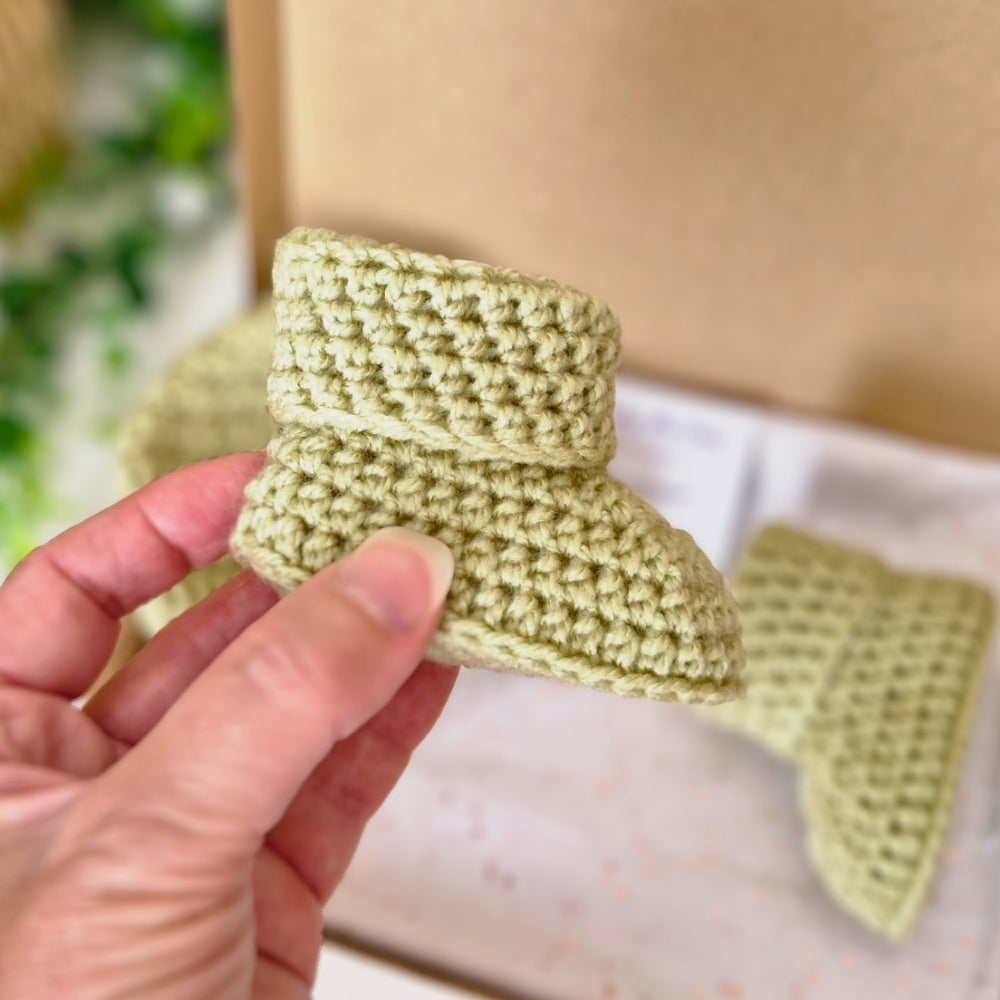 A hand holding a small handmade crochet bootie in a light sage green color, with another matching bootie and a cardboard box blurred in the background.