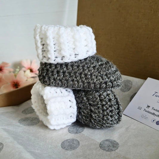 A pair of crocheted booties, white and gray, stacked on a surface with a blurred background.