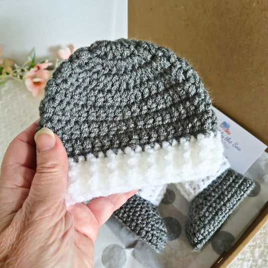 Hand holding a gray and white knitted baby hat with matching booties on a cardboard box.