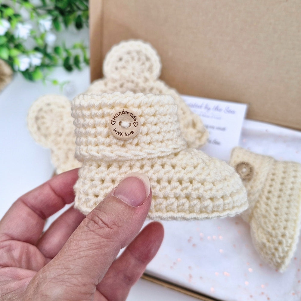 A hand holds a cream-colored crocheted baby bootie and small wooden button that reads 'handmade baby love'. In the background, a kraft gift box and a matching cream crocheted bootie are visible.