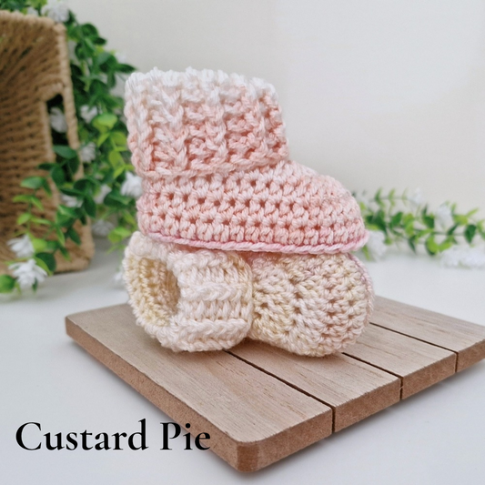 Three crocheted bootie-style baby shoes in soft pastel shades labeled 'Custard Pie,' stacked on a small wooden board with decorative greenery in the background.