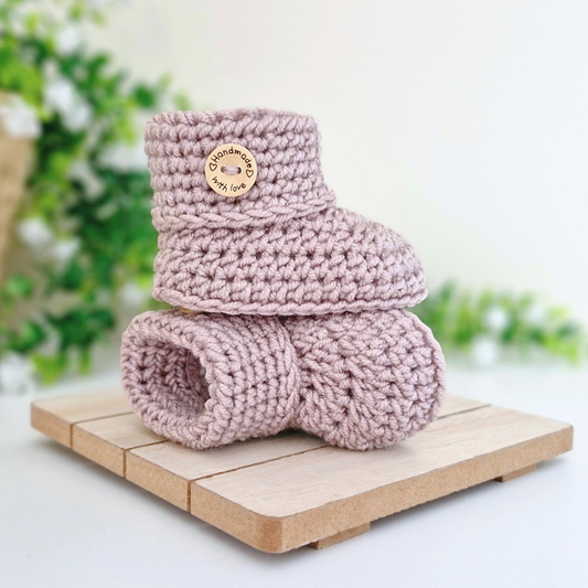 A pair of dusty pink crocheted baby booties stacked on a small wooden block, with a wooden button that reads 'handmade with love'.
