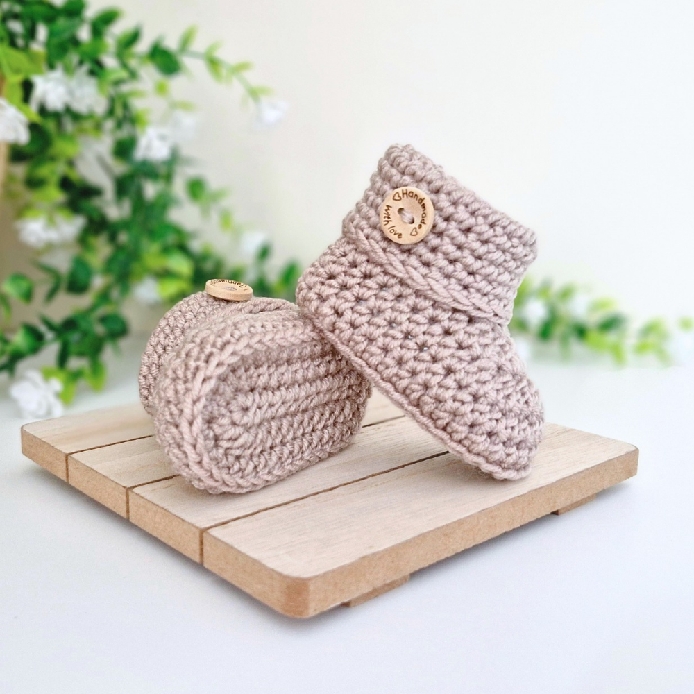 A pair of crochet baby booties in a light taupe-beige color, displayed on a small wooden block with a soft-focus background of green leaves and white flowers.