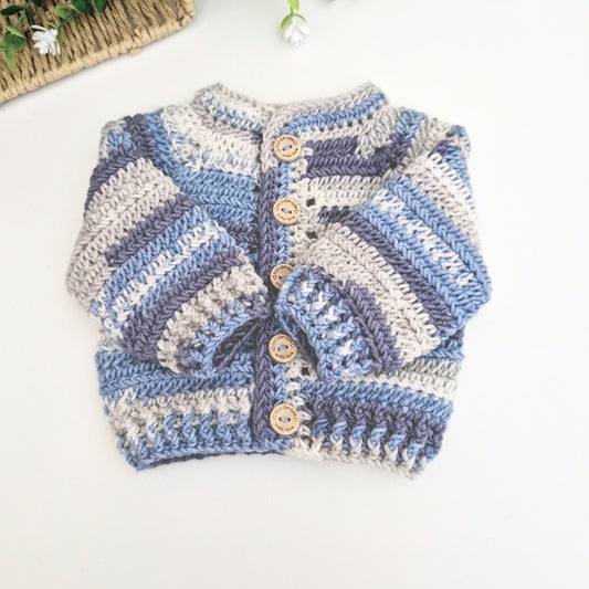 A handmade crocheted baby cardigan with horizontal stripes in light blue, medium blue, and cream, featuring wooden buttons down the front, laid flat on a white surface.