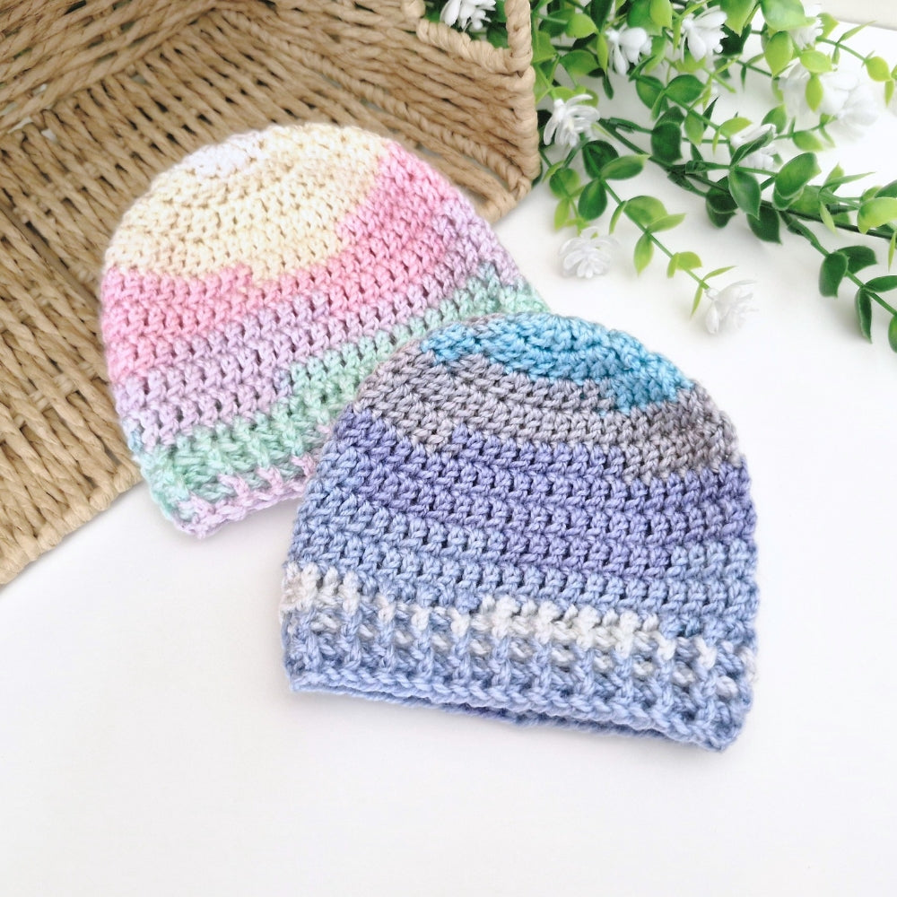 Two crocheted baby beanie hats displayed on a white surface with decorative props. One hat has pastel stripes in cream, pink, mint green, and lavender.