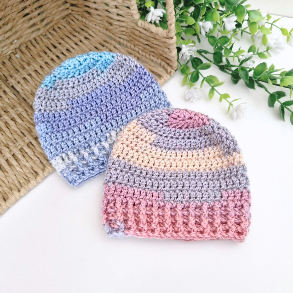 Two crocheted knit beanies displayed on a white surface with a woven basket and greenery in the background. One beanie is blue and lavender, and the other is pink, peach, and gray.