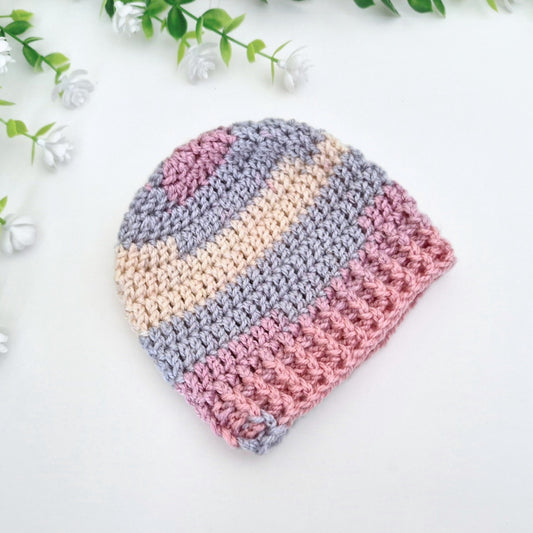 A handmade crocheted beanie hat with horizontal stripes in dusty pink, light blue-gray, and cream, styled on a white surface with decorative green leaves and small white flowers.