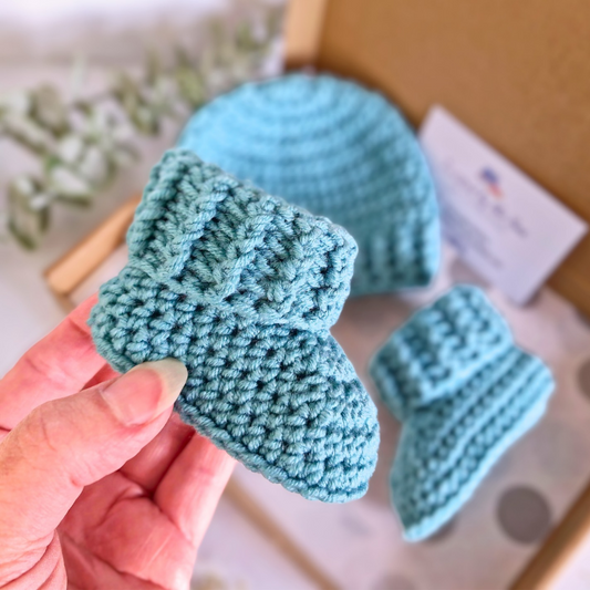 Hand holding a pair of blue knitted baby booties with a matching hat in the background.