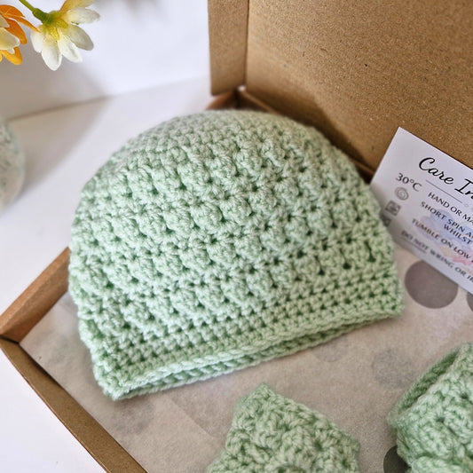 Green crocheted baby hat and booties in a box with care instructions.