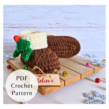 Brown crocheted baby booties with white cuffs and green holly decoration on wooden board with Christmas bells