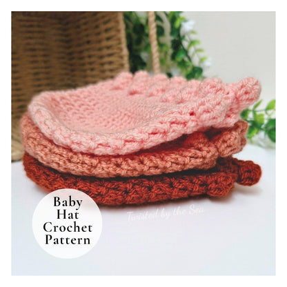 Three crocheted baby hats stacked on a white surface, with a white circular label reading 'Baby Hat Crochet Pattern' and a faint 'Twisted by the Sea' watermark. The hats are in soft pink, dusty rose, and rust orange tones, with a woven basket and green leaves blurred in the background.