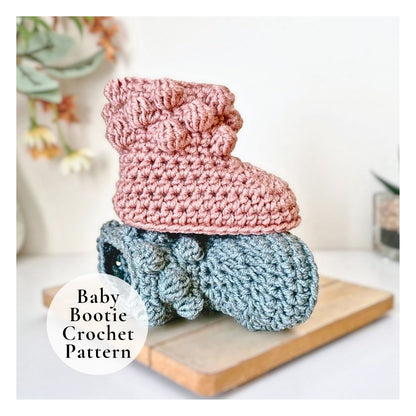 Two crocheted baby booties, one pink and one blue, on a wooden board with a blurred plant in the background.