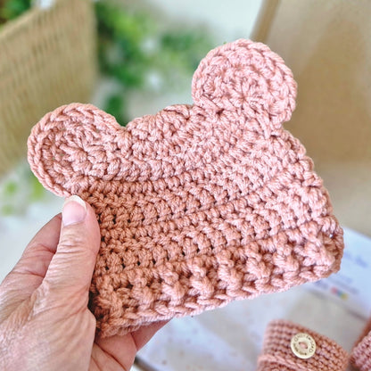 Milky tea coloured crochet baby hat with ears held by a hand against a blurred background