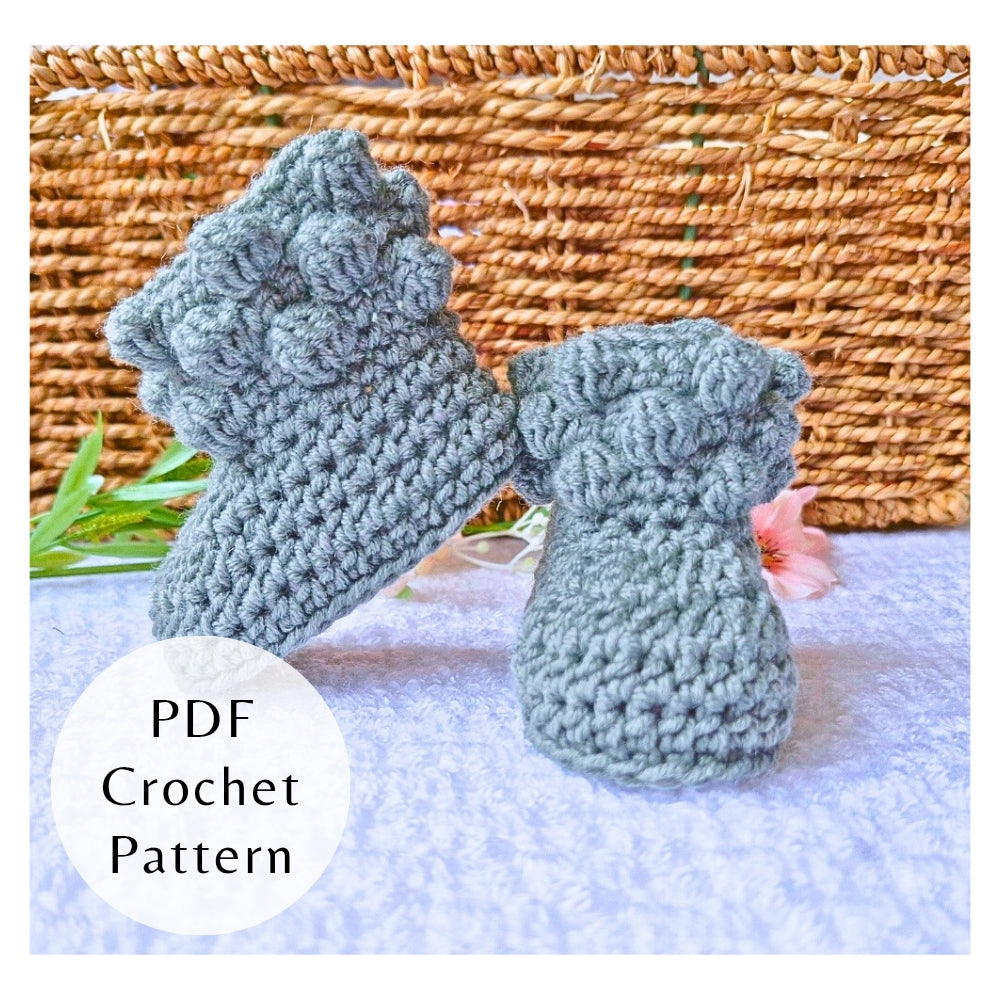 Pair of crocheted baby booties on a textured surface. PDF Crochet Pattern by Twisted by the Sea, pictured in blue merino wool.