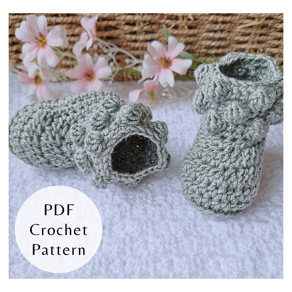 Handmade crochet pattern for bobble baby booties by Twisted by the Sea. Digital PDF download featuring US crochet terms and step-by-step photos. Crocheted baby booties on a white surface with pink flowers and a woven basket in the background.