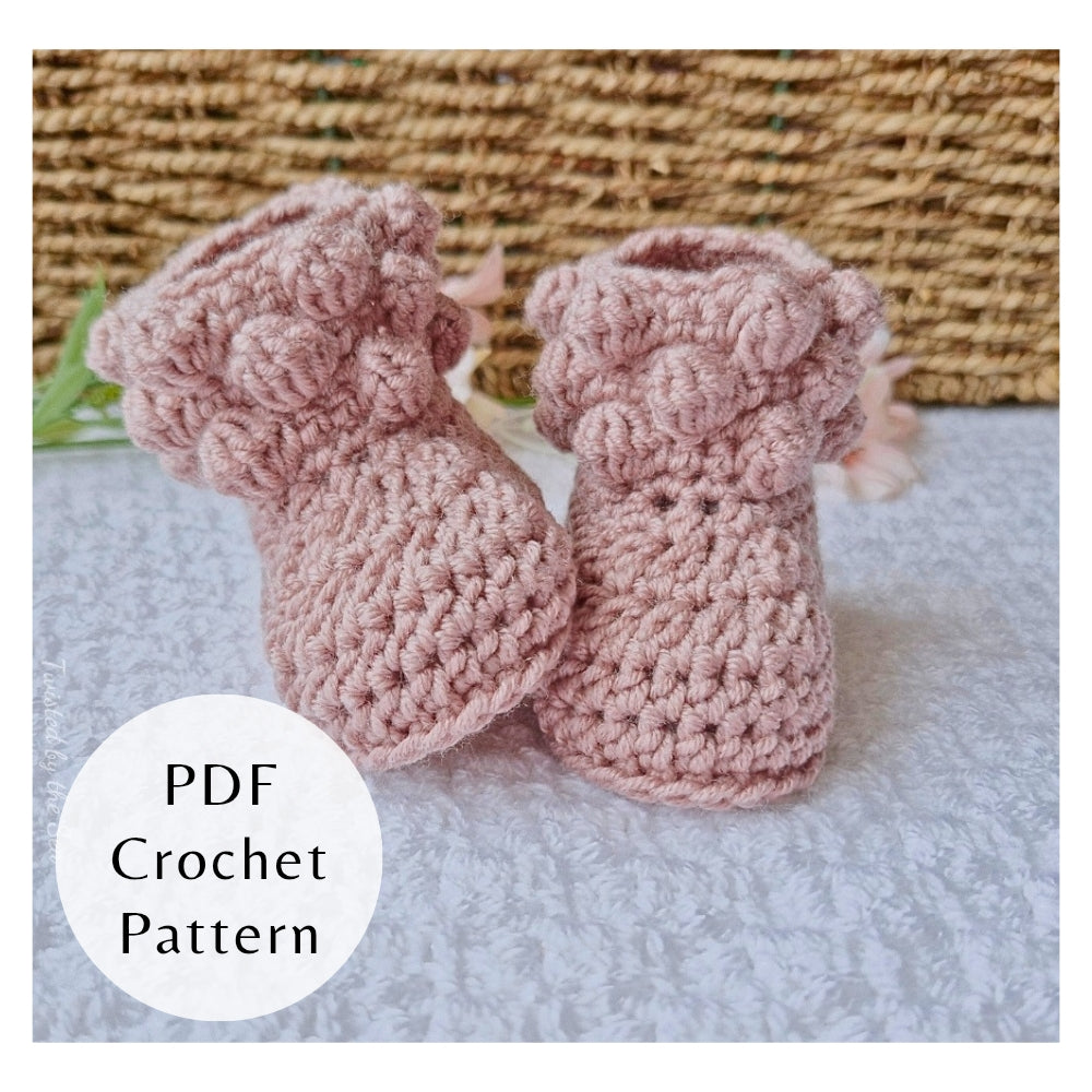 Pair of pink crochet baby booties on a textured surface, PDF Crochet Pattern for confident beginners by Twisted by the Sea. 