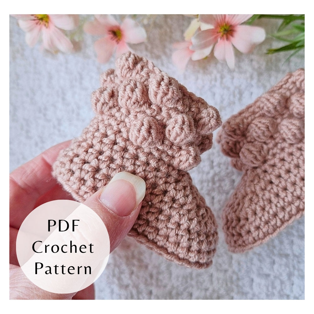 Crocheted bobble baby booties held by a hand with flowers in the background, a PDF crochet pattern by Twisted by the Sea. 