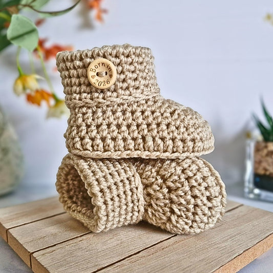 Crochet baby booties in parchment with born in 2026 wooden buttons, on a wooden stand.