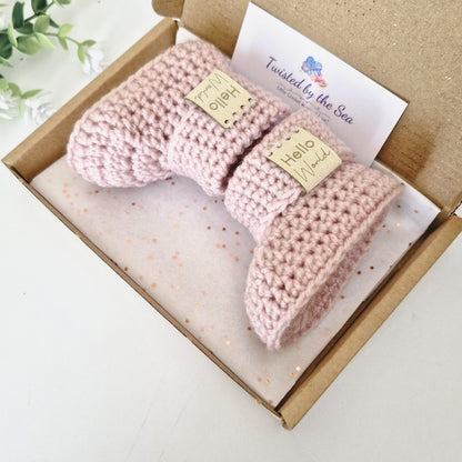 Pink baby booties in a box with a 'Twisted by the Sea' label on a white background