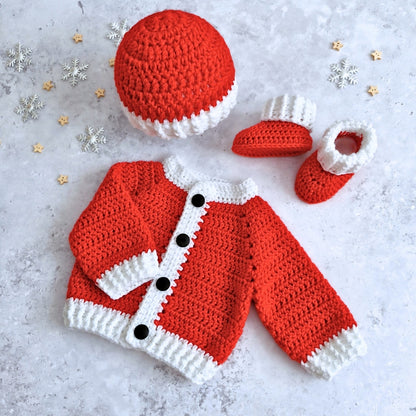 A crocheted baby outfit set arranged on a light stone surface, including a Santa hat, cardigan, booties, and small decorative snowflakes and stars.