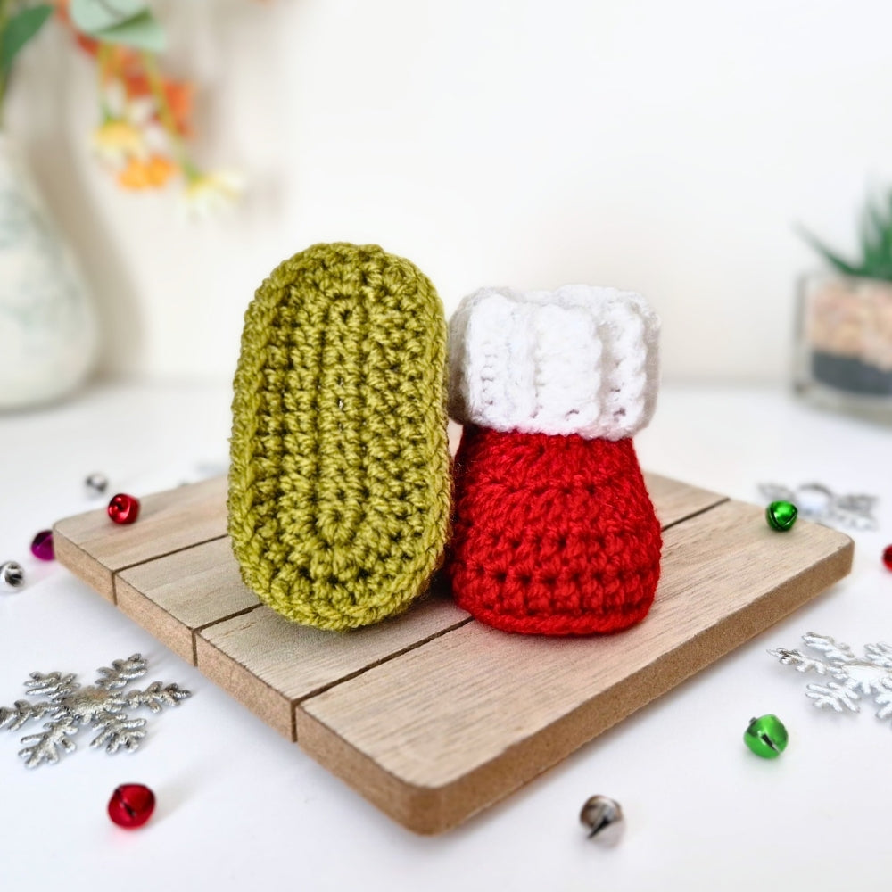 Two crocheted christmas baby booties, one mistletoe green and one red with a white cuff, displayed on a small wooden board with festive decorations.