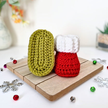 Two crocheted christmas baby booties, one mistletoe green and one red with a white cuff, displayed on a small wooden board with festive decorations.