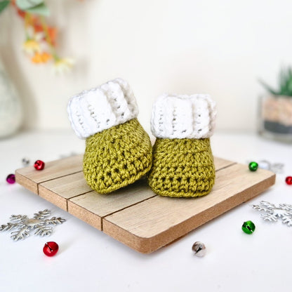 A pair of handmade crocheted baby booties styled on a small wooden tray, decorated with red and green holiday ornaments and silver snowflake cutouts on a white surface.