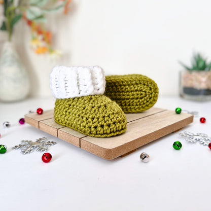 A pair of handmade crocheted baby booties in mistletoe green with white cuff trim, displayed on a small wooden board with Christmas decorations in a soft-focus indoor setting.