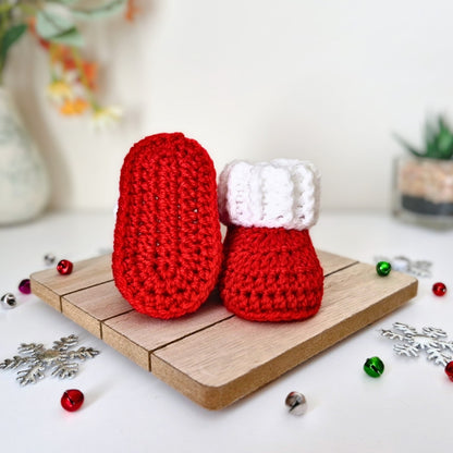 A pair of red and white crocheted baby booties styled on a small wooden board with christmas decorations in the background.