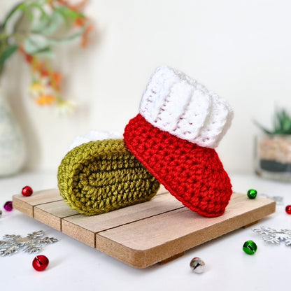 A pair of handmade crocheted booties styled on a small wooden board with christmas decorations. One bootie is bright red with a white cuff, and the other is mistletoe green.