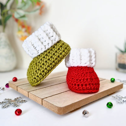 Two crocheted baby booties styled like Christmas stockings, one in mistletoe green and white, and one in red and white, displayed on a wooden board with Christmas decorations.