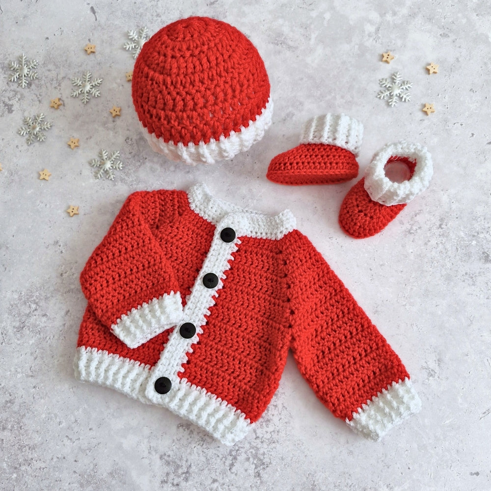A crocheted baby festive outfit set laid flat on a light textured surface, including a red and white Santa hat, cardigan with black buttons, and matching booties. Small silver and gold snowflake decorations are scattered around.