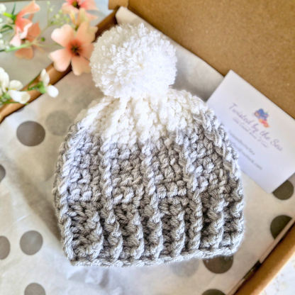 Crochet baby hat with a pom-pom on a polka dot surface, with a brand card in the background.