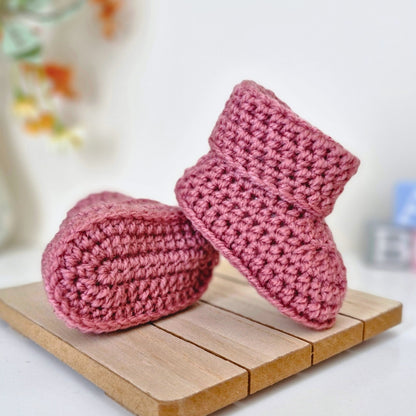 Cinder rose deep pink Pair of crochet baby booties on a wooden surface with a blurred background