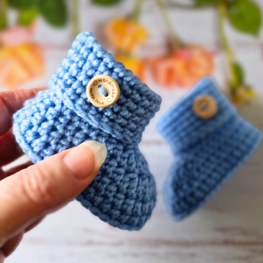 Blue crochet baby booties with a button held by a hand against a blurred background.