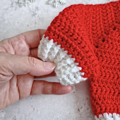 A close-up of a hand holding a red and white crocheted item, showing the textured knit pattern and the white trim against a light gray background.
