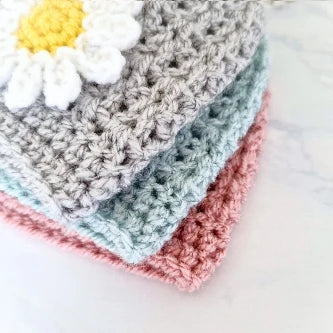 Close-up of textured crochet fabric squares in pink, blue, and gray with a white and yellow daisy applique
