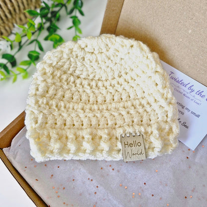 Cream crocheted baby hat with 'Hello World' tag in a box on a light background
