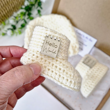 Cream crocheted baby booties with 'Hello World' tag held by a hand, with more booties in the background.