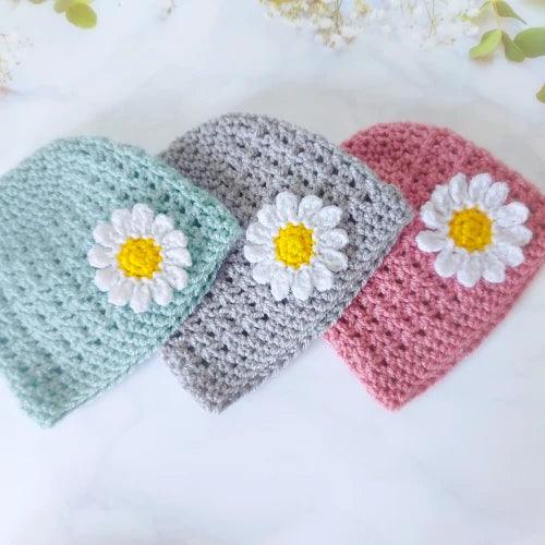 Three crocheted baby hats in duck egg blue, silver gray, and pink with white daisy appliques on a white surface