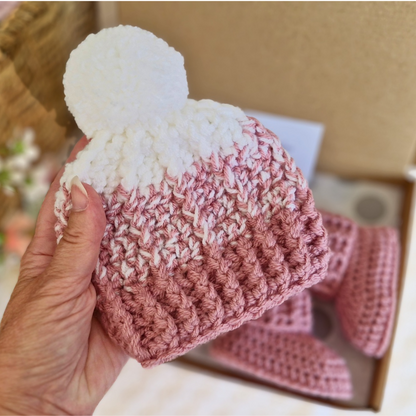 Hand holding a pink and white crocheted beanie with a matching pair of booties in the background.