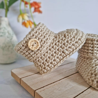 Beige crochet baby booties with a born in 2026 button on a wooden surface, flowers in the background
